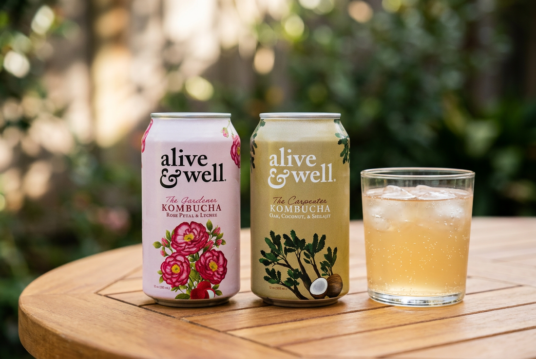 Alive & Well Rose Petal and Lychee and Oak and Coconut kombucha cans beside an iced drink on a wooden table with a garden backdrop.
