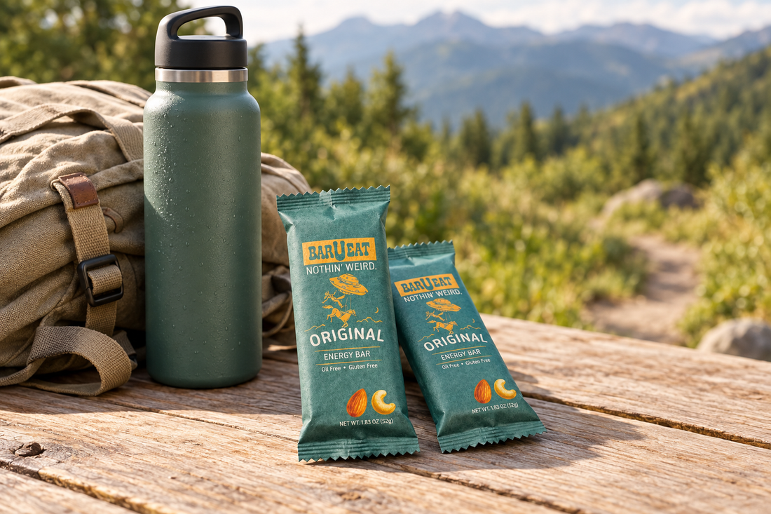 BAR-U-EAT Original energy bars beside a water bottle and canvas daypack on a wooden picnic table, with mountain greenery softly blurred behind them.