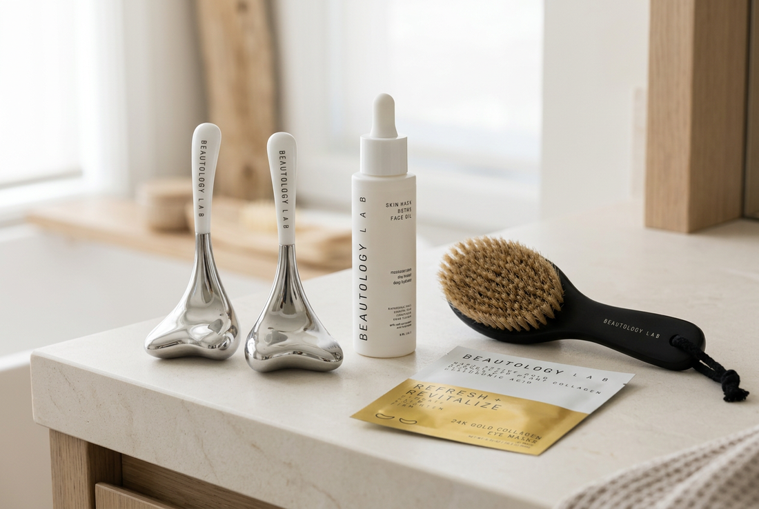 BEAUTOLOGY LAB silver cryo tools, face oil bottle, black dry brush, and gold eye-mask packet arranged on an ivory stone vanity.