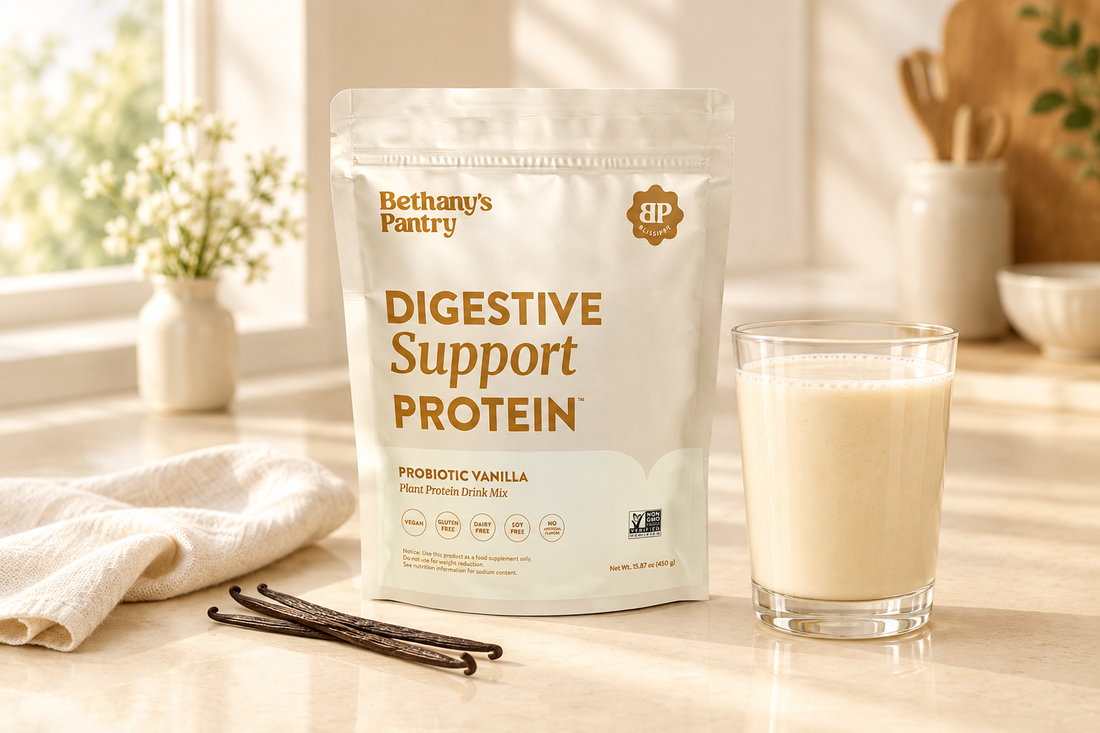 Bethany’s Pantry Digestive Support Protein Probiotic Vanilla pouch beside a glass of vanilla-colored drink, vanilla beans, and ivory linen on a cream countertop.