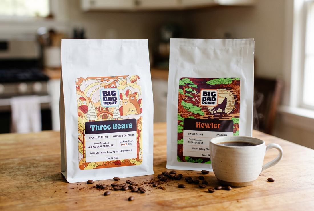 Big Bad Decaf Three Bears and Howler coffee bags beside a ceramic cup of coffee and coffee beans on a wooden table.