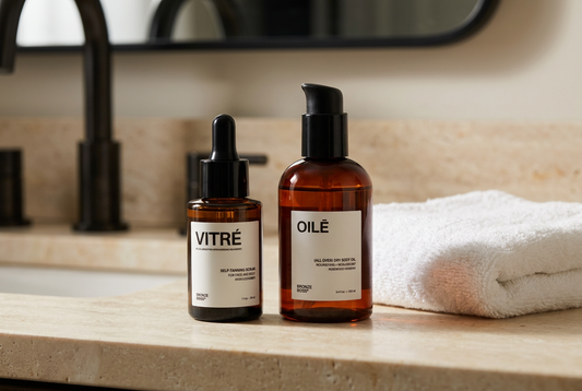 Bronze Boss Vitré self-tanning serum and Oilē dry body oil on an ivory stone vanity beside a folded white towel.