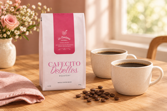 Cafecito Debelles Roastery La Santana coffee package beside two cream cups of coffee, roasted beans, and blush linen on a warmly lit wooden table.