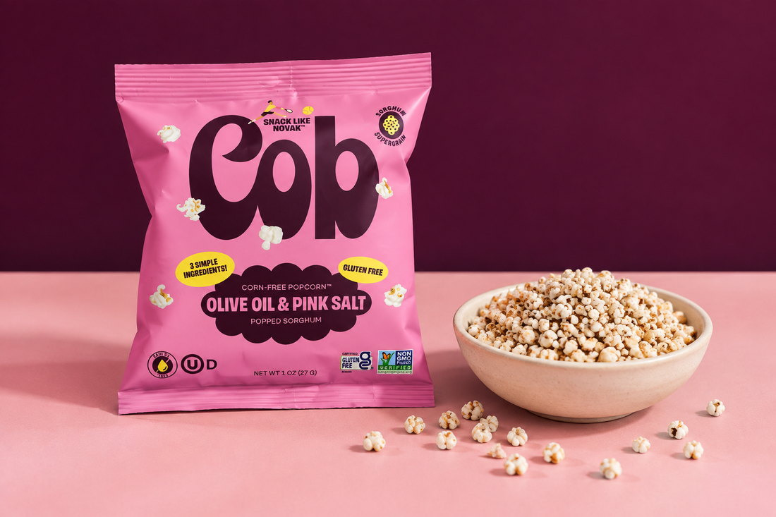 Cob Olive Oil Pink Salt popped-sorghum bag beside a cream bowl of popped sorghum on a pink tabletop against a deep-plum background.