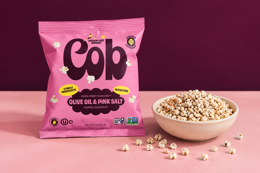 Cob Olive Oil Pink Salt popped-sorghum bag beside a cream bowl of popped sorghum on a pink tabletop against a deep-plum background.