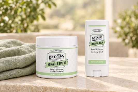Dr. Doug’s Original Miracle Balm jar and balm stick beside sage-green linen on a warmly lit cream stone surface. ⠋