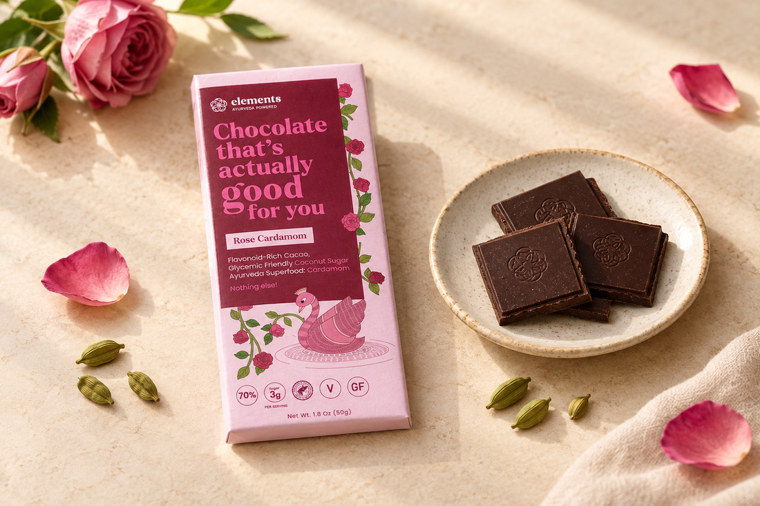 Elements Truffles Rose Cardamom chocolate packaging beside dark chocolate squares, rose petals, and cardamom pods on a cream tabletop.