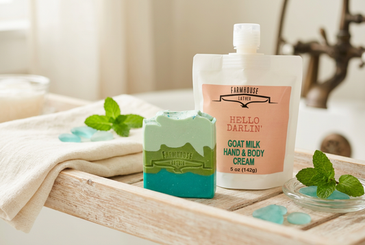 Farmhouse Lather Suns Up Serenade turquoise and green soap beside a Hello Darlin’ goat-milk hand-and-body cream pouch on a wooden tray with ivory linen.