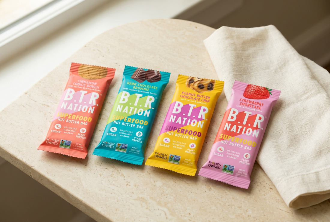 Four B.T.R. NATION Superfood bars in coral, teal, yellow, and pink wrappers beside ivory linen on a cream stone tabletop.