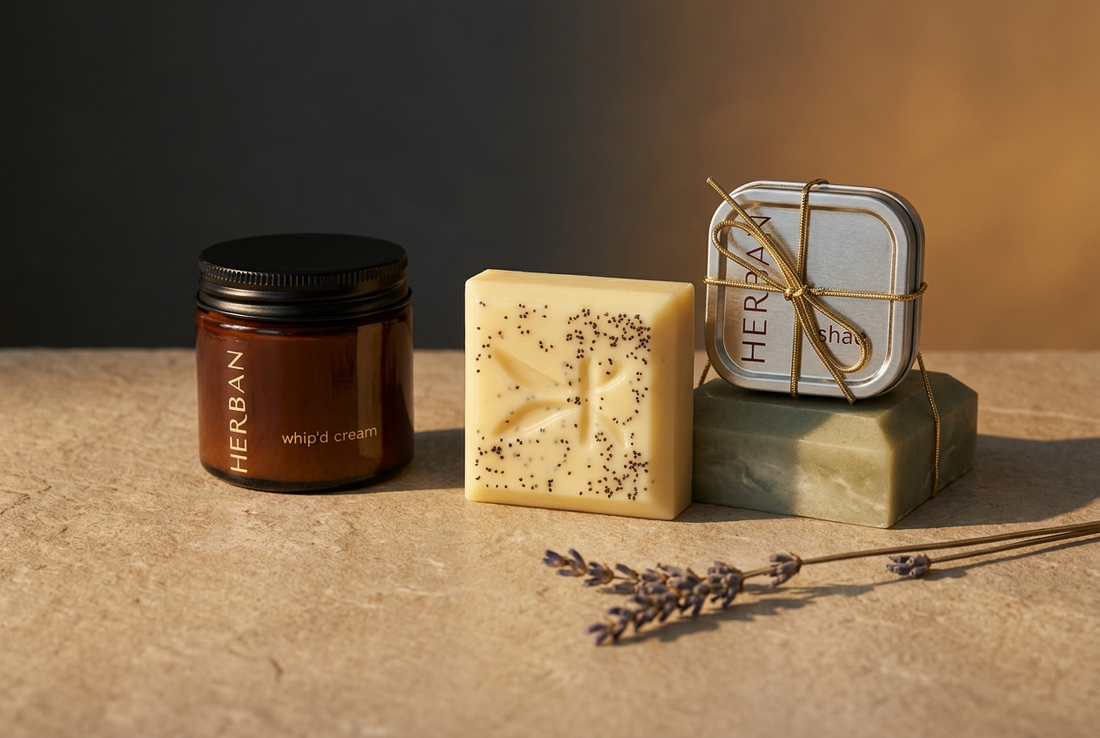Herban Body Care amber Whip’d Cream jar, botanical soap bar, and silver Shae tin arranged on warm oat-colored stone against an amber and charcoal background.