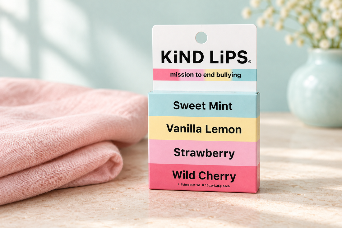 Kind Lips four-flavor variety box with pastel color bands beside pale-pink linen on a cream stone surface with a soft aqua background.