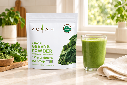 KOYAH Organic Greens Powder pouch beside a glass of green smoothie and ivory linen on a bright cream kitchen countertop.