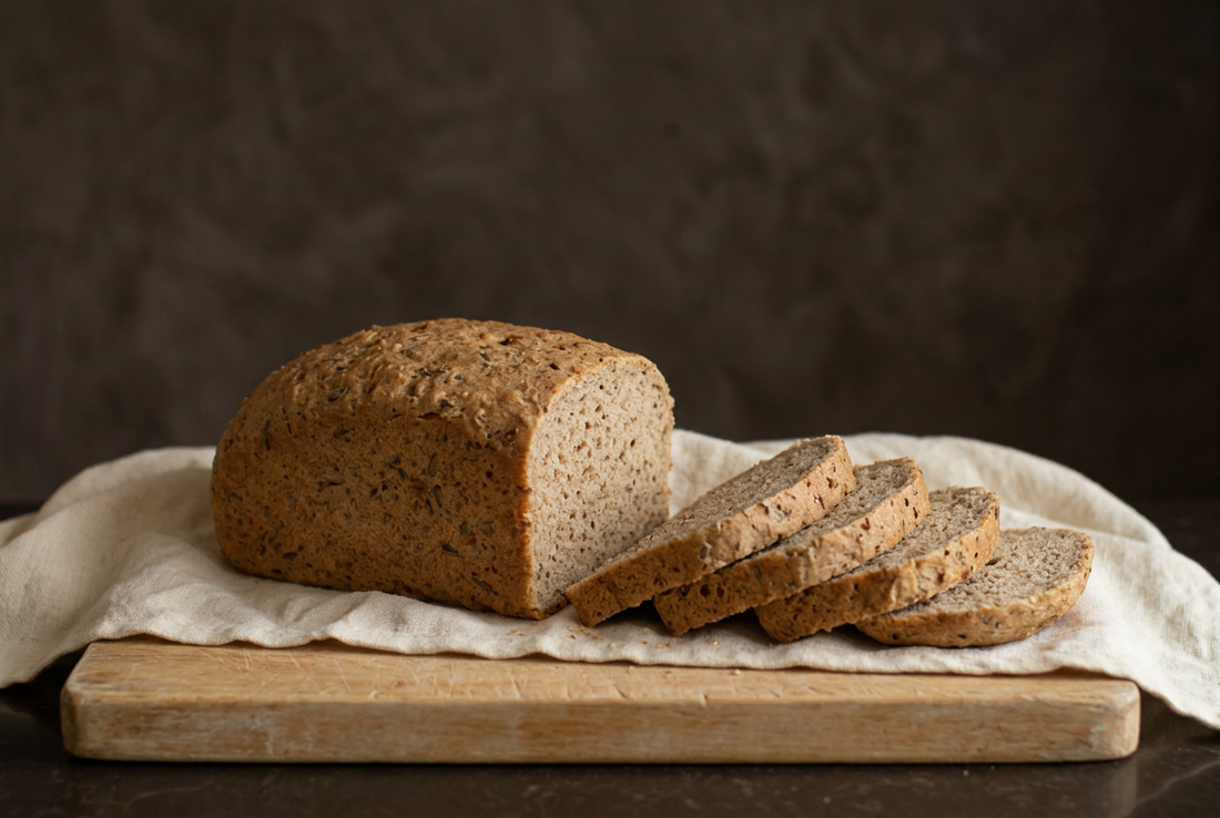 Leaven & Love gluten-free sourdough loaf and slices on a wooden bread board with ivory linen and a warm charcoal background.