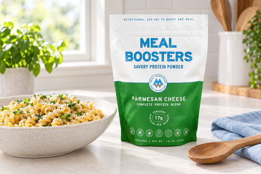 Meal Boosters Parmesan Cheese savory protein powder pouch beside a bowl of pasta, a wooden spoon, and pale-blue linen on a bright kitchen counter.
