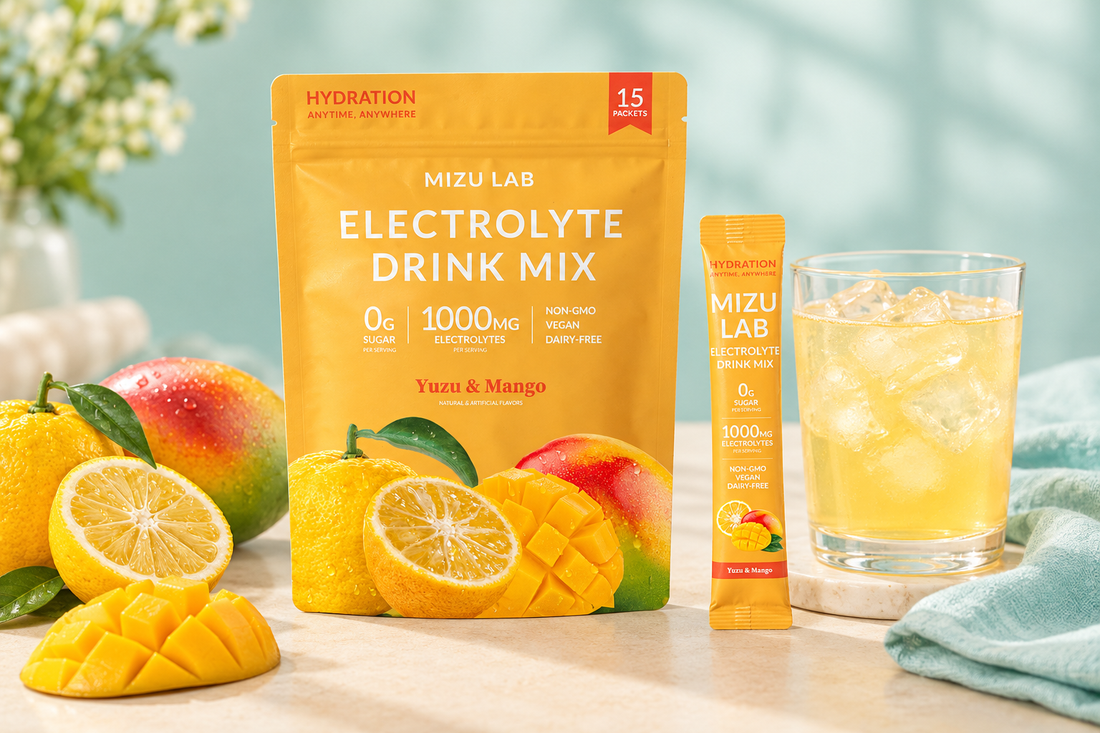 Mizu Lab Yuzu and Mango electrolyte drink mix pouch and stick beside an iced drink, mango pieces, citrus, and pale-aqua linen on a cream tabletop.