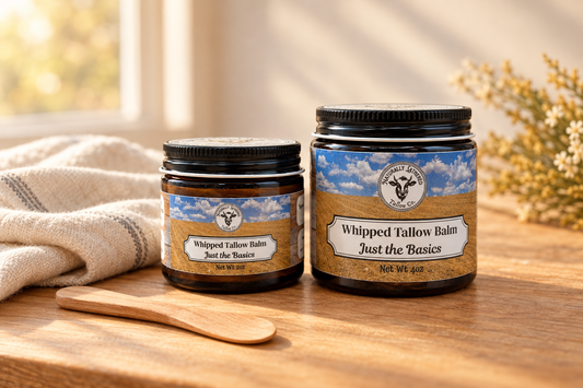 Naturally Lathered Tallow Co. Just the Basics whipped tallow balm jars beside cream linen and a wooden cosmetic spatula on a warmly lit wooden surface.