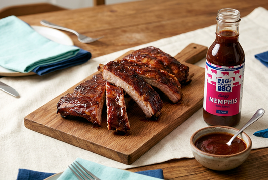 Pig of the Month BBQ glazed baby back ribs on a wooden board beside a Tangy Memphis BBQ sauce bottle and a turquoise napkin.