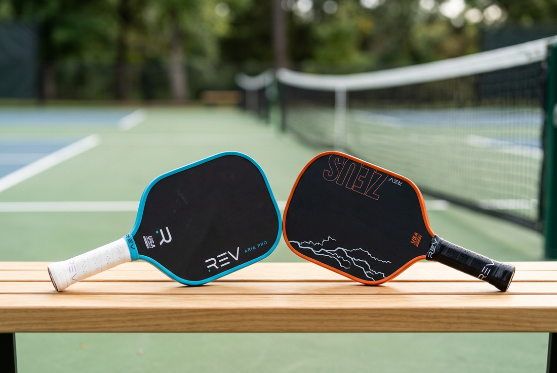 REV Pickleball ARIA PRO paddle with cyan trim and ZEUS paddle with orange trim resting on a bench beside a pickleball court.