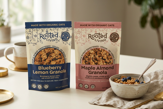 Rooted Living Blueberry Lemon and Maple Almond granola bags beside a ceramic bowl of granola and blueberries on an ivory breakfast table.