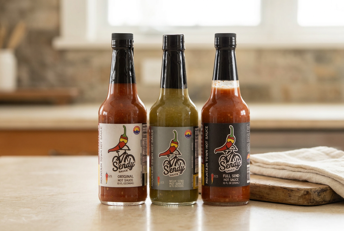 Sendy Sauce Original, Weak Send, and Full Send bottles beside ivory linen and a wooden board on a cream kitchen counter.