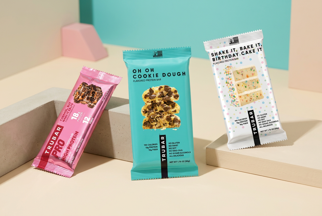 TRUBAR Cookie Dough, Birthday Cake, and pink PRO Caramel protein bars arranged on a cream surface against a turquoise background with pink accents.