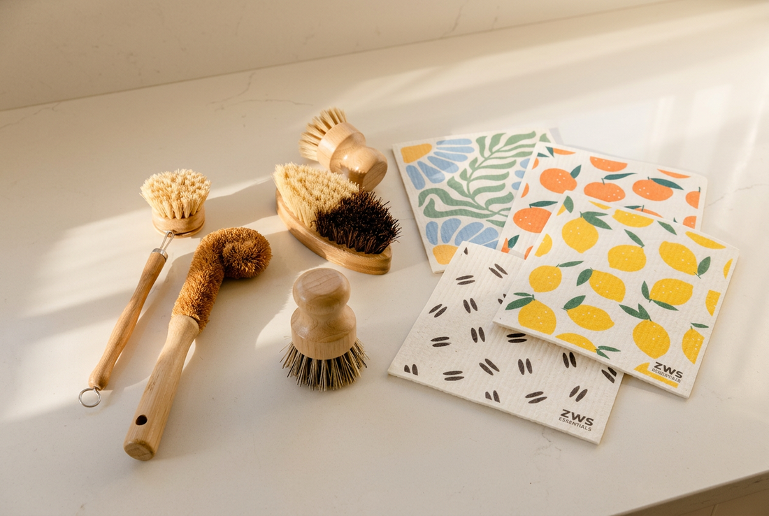 Wooden dish brushes and patterned ZWS Essentials Swedish dishcloths with blue flowers, oranges, lemons, and seed designs on an ivory kitchen counter.