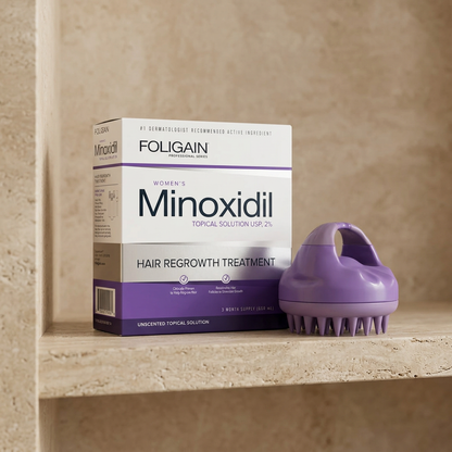 Boxed FOLIGAIN women's 2% minoxidil and purple scalp massager on a warm neutral stone bathroom shelf.