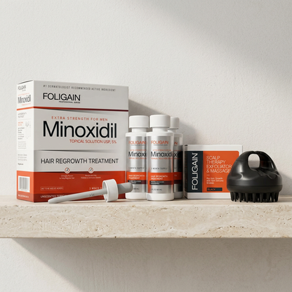 FOLIGAIN men's 5% minoxidil three-month supply and black silicone scalp massager with their packaging arranged on a pale stone shelf.